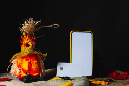 Credit card on a cell phone with a yellow case and the screen on, both standing next to an ornate Halloween pumpkin, on sackcloth and surrounded by toy spiders and fallen leaves next to pine cones.の写真素材