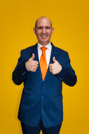Portrait of a smiling bald Caucasian adult male, dressed in blue suit, white shirt and orange tie, with both thumbs up looking at camera. The background is yellow.の写真素材
