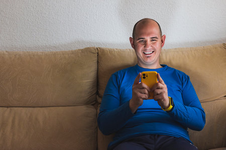 A smiling bald Caucasian man wearing a blue long-sleeved T-shirt is using a cell phone sitting on a...