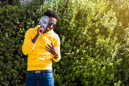 A smiling young African man dressed in a yellow sweatshirt, blue jeans and sunglasses holding some bills next to his face while waving his fingers with his other hand.の写真素材