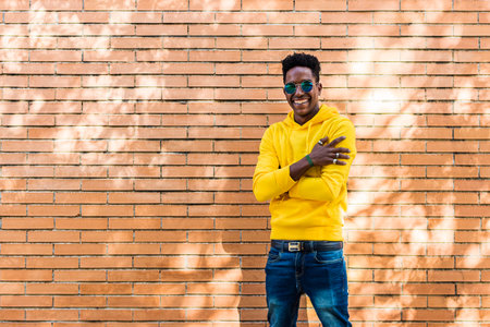 Portrait of a smiling young African man wearing a yellow hooded sweatshirt, jeans and sunglasses in front of a brick wall. The guy is wearing rings on his fingers.の写真素材