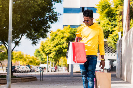 A young African man dressed in a yellow sweatshirt, jeans and sunglasses is walking down the street holding shopping bags while looking at his cell phone screen.の写真素材