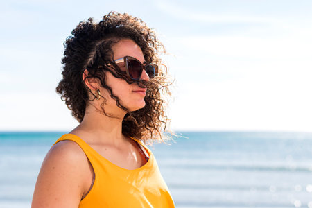 Side view of female with curly hair in sunglasses looking away against serene sea in sunlightの写真素材