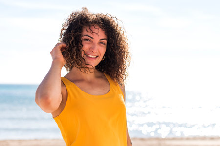 Happy curly female looking at camera with toothy smile and touching hair on coast against blurred sea in summer dayの写真素材