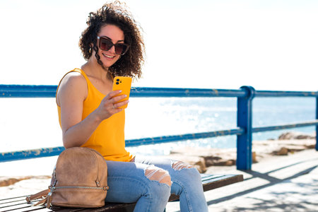 Cheerful young curly haired female in sunglasses and casual outfit smiling while sitting on wooden bench with backpack and messaging via smartphone against waving sea during summer tripの写真素材