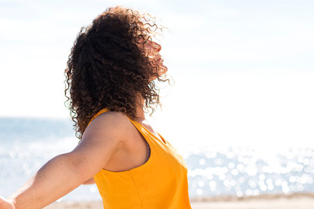 Side view of satisfied curly brunette spreading arms in front of blurred rippling sea on sunny dayの写真素材