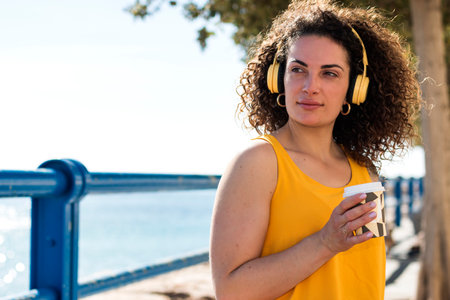 Pensive young female in casual top with curly hair looking away while listening to music in...