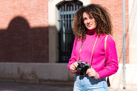 Young curly haired woman in casual outfit standing on town street against brick building with vintage photo camera while exploring cityの写真素材