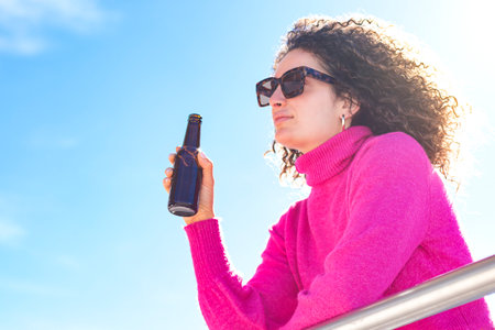 Low angle of young curly haired female in sunglasses and pink turtleneck leaning on metal railing while drinking beer against blue sky with sunlightの写真素材