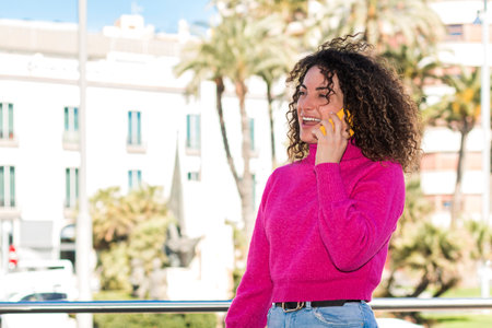 A young beautiful caucasian woman with curly hair wearing a pink jersey is talking with someone with her mobile phone while she is laughing, she is in the street.の写真素材