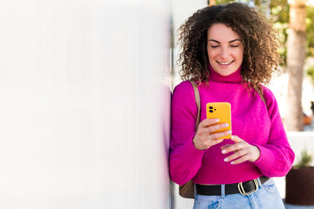 A beautiful young caucasian woman with curly hair touching her mobile phone screen while is smiling leaning to a white wallの写真素材