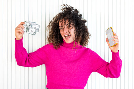 Young caucasian woman with curly hair is happy while holding with a hand some 50 dollar bills and a smartphone in the other hand in front of a wall.の写真素材