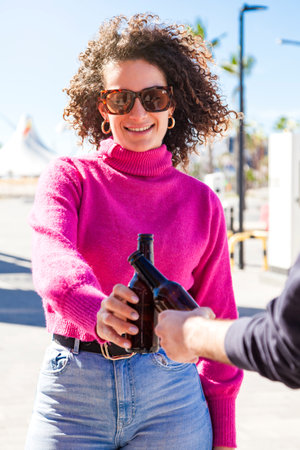 Curly hair young caucasian woman making a toast with a crop person in the street holding two beer bottles on a sunny day.の写真素材