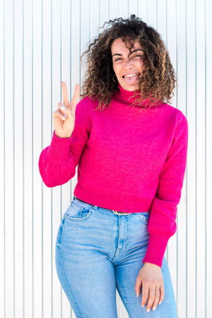 Joyful adult woman in pink sweater standing near white wall with tongue out and showing two fingers gesture while looking at cameraの写真素材