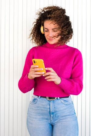 Positive curly haired female in casual clothes and headphones standing near striped wall and...