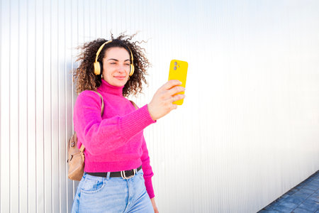 Young caucasian woman with curly hair taking a self portrait with her smartphone while leaning to a wall and listening to music with headphones.の写真素材