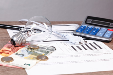 Over a wooden table an electricity bill, light bulb, euro money, calculator, pen, cord with european plug.の写真素材