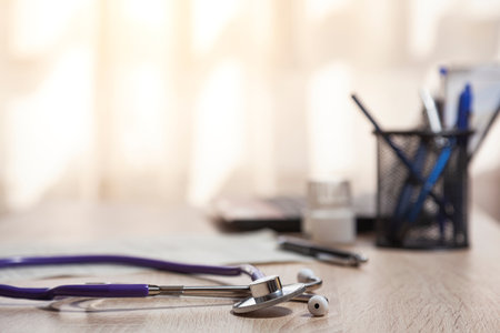 Close-up of a stethoscope over a wooden table where, out of focus, a pen pot, paper, medicines and a laptop. There are a window on background.の写真素材