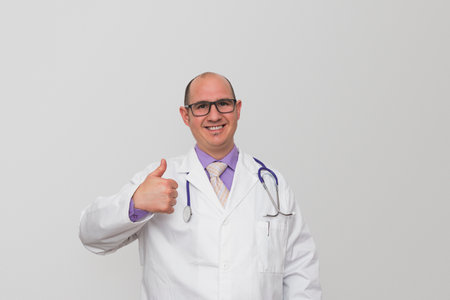 A bald male smiling caucasian doctor with eyeglasses wearing a white coat over a purple shirt have on thumb up gesturing. Background is light grey.の写真素材