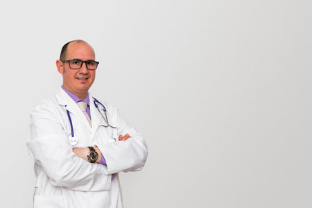 A bald male smiling caucasian doctor with eyeglasses wearing a white coat over a purple shirt with arms crossed. Background is light grey.の写真素材