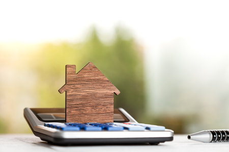 Close up shot of wooden silhouette in the shape of a house standing on a calculator next to a pen on top of a table in front of a window.の写真素材