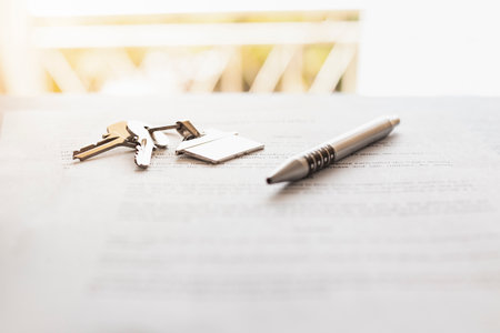 Close-up of a house-shaped key ring with keys and a pen on a house purchase contract on a table in front of a window.の写真素材