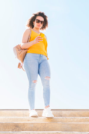 Young beautiful caucasian woman with curly hair wearing jeans, yellow blouse, backpack and sunglasses is going down a stairs while using her smartphone.の写真素材