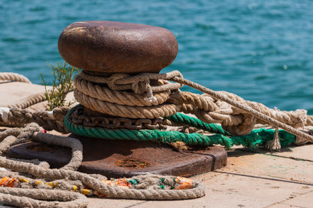 Close-up of a rusty metal mooring wrapped in thick ropes and chains on the pavement of the harbor by the sea.の写真素材