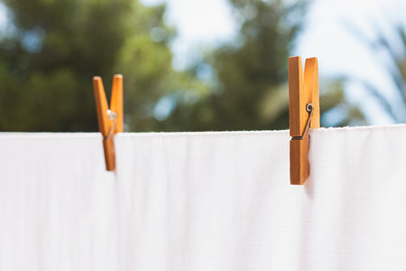 White cloth placed on clothesline while drying in sunlight and fastened with peg of two wooden levers attached with metal spring against blurred green trees and blue skyの写真素材