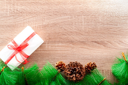 Seen from above, one border with part of a pine tree, pine cones and a gift box with a red ribbon over a wooden table.の写真素材