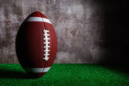 An American football ball standing over green grass in front of a gray wall.の写真素材