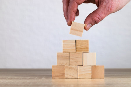 Close-up of a hand stacking a wooden block on top of a pyramid made of identical blocks against a...