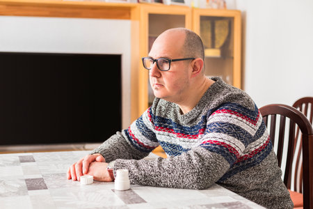 A bald adult male sitting at a table next to a medicine bottle with a sad and worried look towards the horizon.の写真素材
