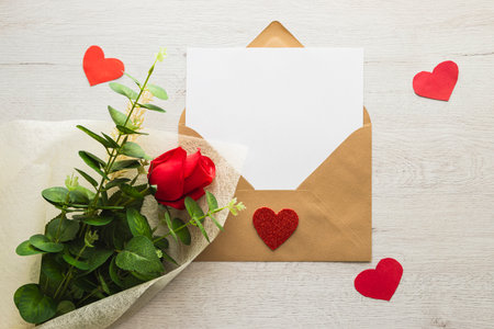 An open kraft paper envelope with a blank sheet of paper inside, next to a red rose and some hearts...