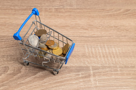 Miniature shopping cart containing various coins on a light wooden background