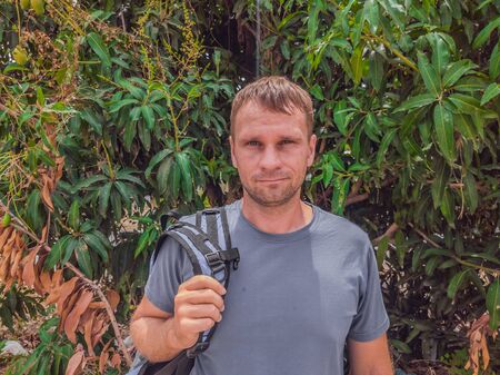 A beautiful young caucasian man in a gray shirt with a backpack near green Mango tree. Travel concept with copy spaceの写真素材