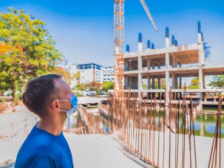 Young man with mouth facial mask from COVID-19 epidemic looking alone at building under construction and crane. Suspension of production due to coronavirus outbreak conceptの写真素材