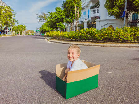 Cute little child boy sitting inside cardboard box on silent street in modern comfortable cottage village. Childhood and moving out conceptの写真素材