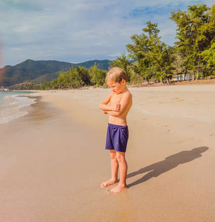 Handsome blond boy in bad mood standing on beach sand near sea in offended and pensive pose. Problem or happy childhood, tourism with child, parent-child relationships, parenting psychology conceptの写真素材