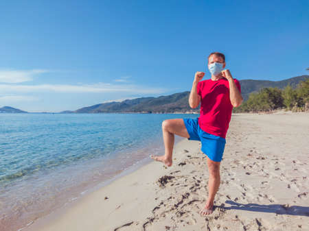 Young muscle man in medical mask perform outdoor workouts in the park near sea during second wave coronavirus Covid 19 pandemic quarantine. Kick training pose, swing arms and legs, sport active lifeの写真素材