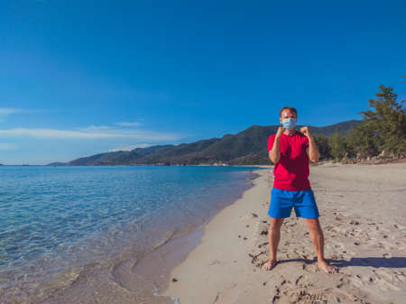 Young muscle man in medical mask perform outdoor workouts in the park near sea during second wave coronavirus Covid 19 pandemic quarantine. Kick training pose, swing arms and legs, sport active lifeの写真素材