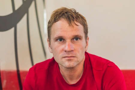 Serious sad handsome blond man in casual style red t-shirt sitting, waiting someone or changes, thinking. Skin without photoshop. Human relationship, difficulties during covid or life problemsの写真素材