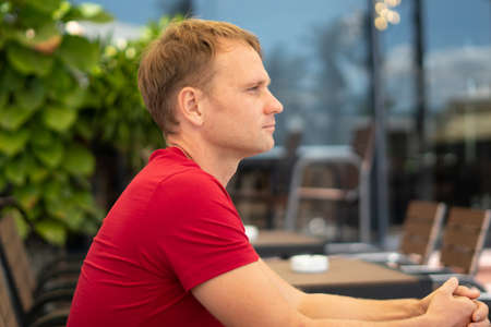 Half side portrait restrained smile handsome young blond man in casual red t-shirt sit in modern cafe, happy look away, waiting someone or met already. Human relationship, good future changes conceptの写真素材