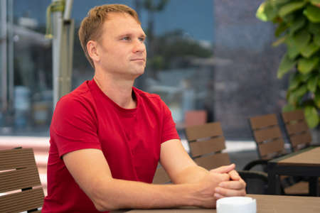 Half side portrait restrained smile handsome young blond man in casual red t-shirt sit in modern...
