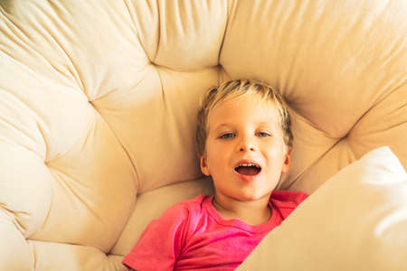 Blond laughing having fun boy open mouth in pink t-shirt sit with pillow in round chair indoor. Family relationship, happy childhood, problem behaviour, educational psychology, child goods producingの写真素材
