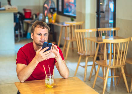 Young serious thoughtful pensive attentive man tense thinking send message by smart phone in red shirt in cafe drinking chia ice tea. Real people expression, life problems solving and communicationの写真素材
