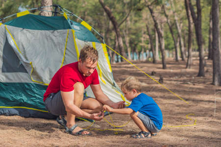 Camping people outdoor lifestyle tourists putting up setting up green grey campsite summer forest near lazur sea. Boy son helps father assembling tent, study metal peg. Natural education of childrenの写真素材