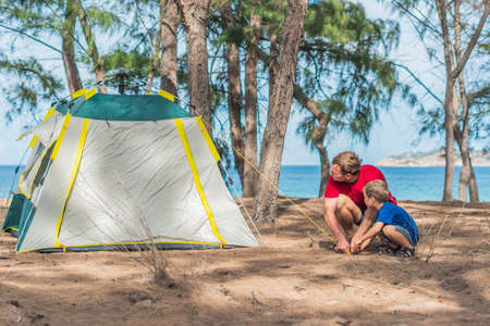 Camping people outdoor lifestyle tourists put up set up green grey campsite summer forest near lazur sea. Boy son helps father study mechanism of modern easy to fold tent. Natural children educationの写真素材