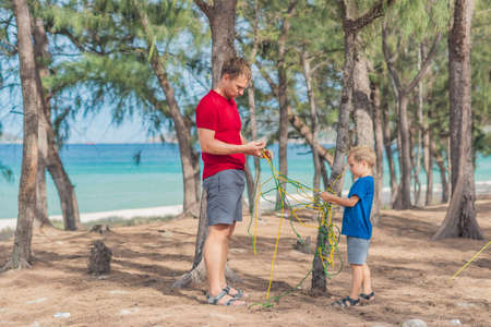 Camping people outdoor lifestyle tourists in summer forest near lazur sea. Blond boy son with father study survival techniques, practice methods of tying rope knots. Natural children educationの写真素材