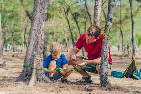 Camping people outdoor lifestyle tourists in summer forest near lazur sea. Blond boy son with father study survival techniques, practice methods of tying rope knots. Natural children educationの写真素材